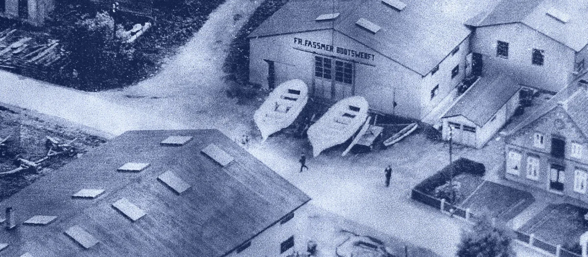 Historic aerial photograph of the Fassmer shipyard in Berne around 1940, showing several workshop buildings, shipyard workers, and two new hulls in front of the hall labeled ‘Fr. Fassmer Bootswerft’.