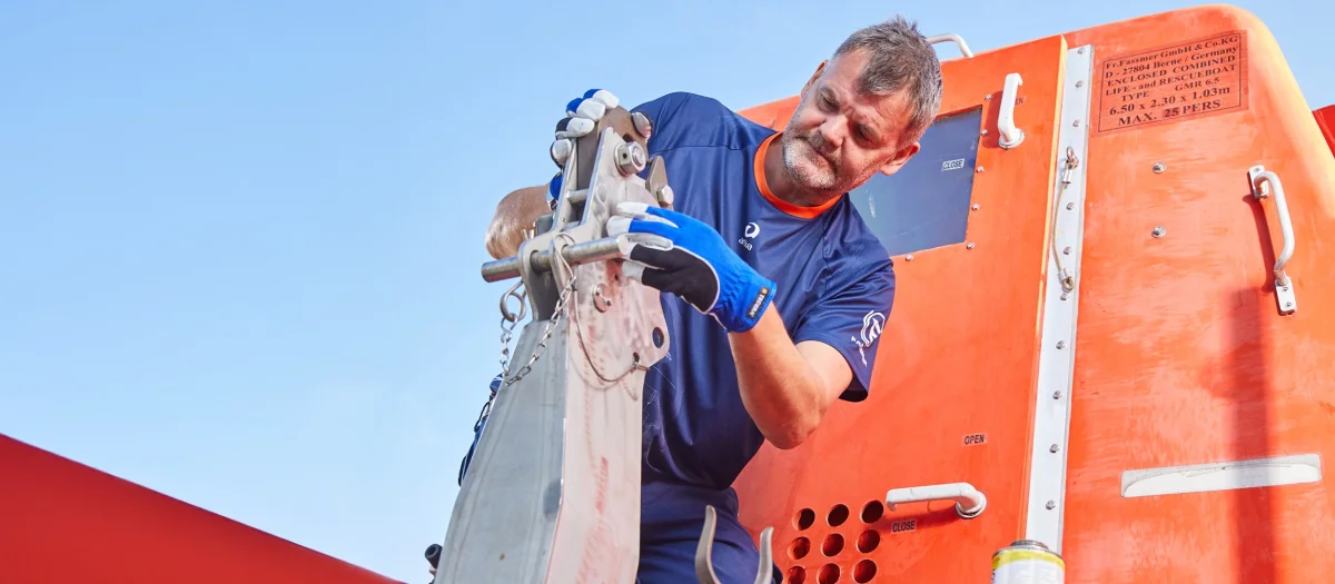 Photo of a Fassmer employee in blue workwear working on a metal component in front of an orange boat hull.