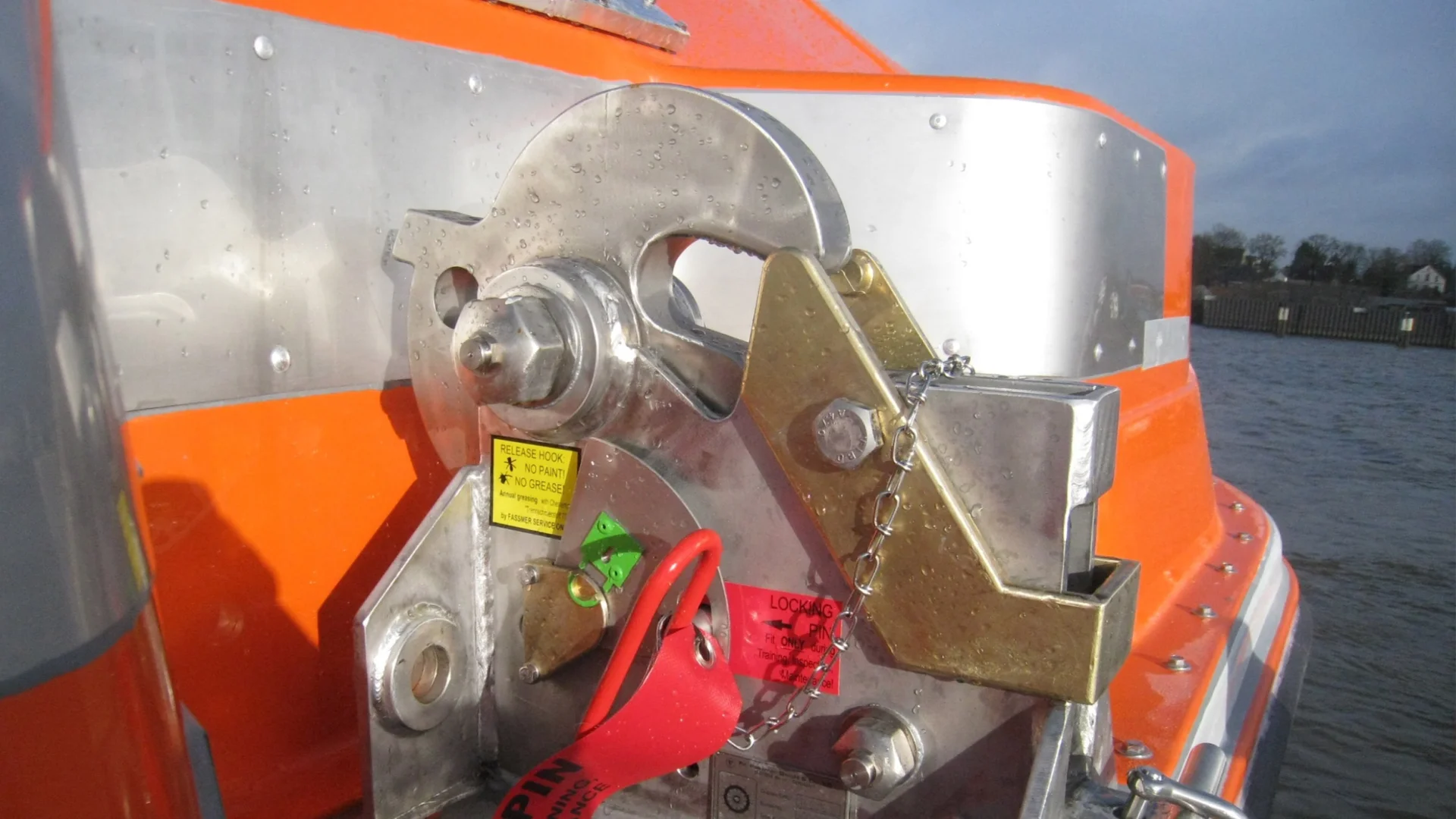 Close-up of a Fassmer original spare part: metallic release and locking mechanism of an orange lifeboat with safety labels.