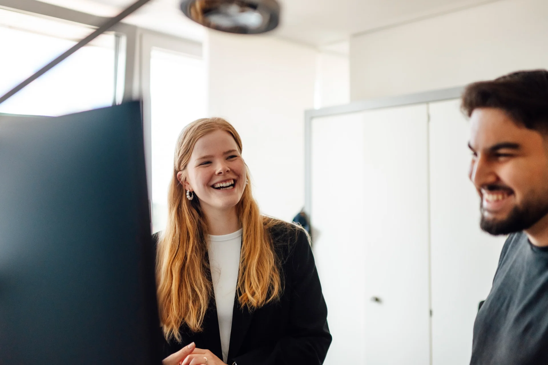 Two young employees at Fassmer laughing together in a modern office. Teamwork and career opportunities in shipbuilding.