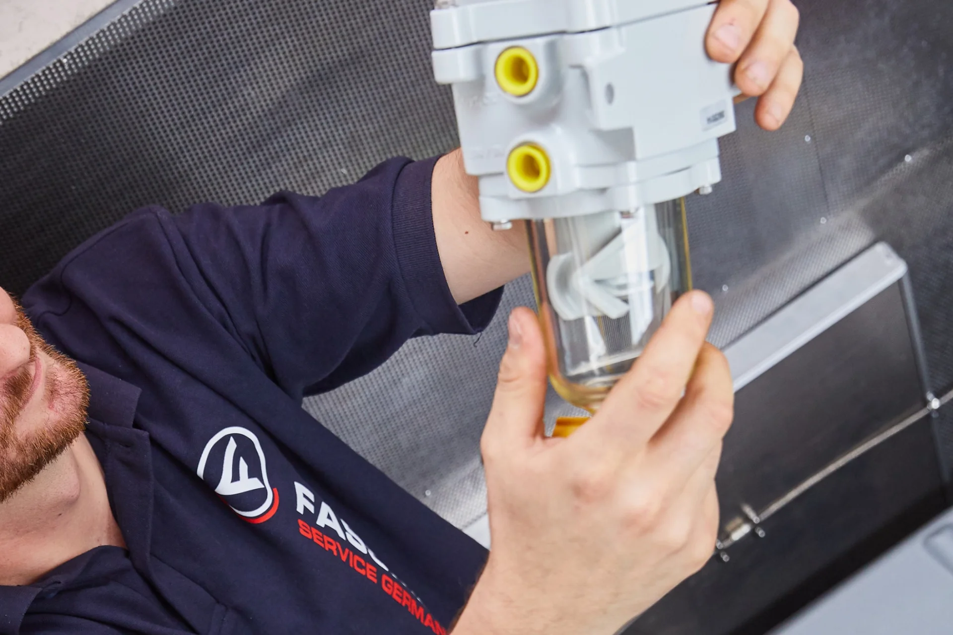 Photo of a Fassmer service technician inspecting a technical spare part with a transparent glass container and yellow connectors.