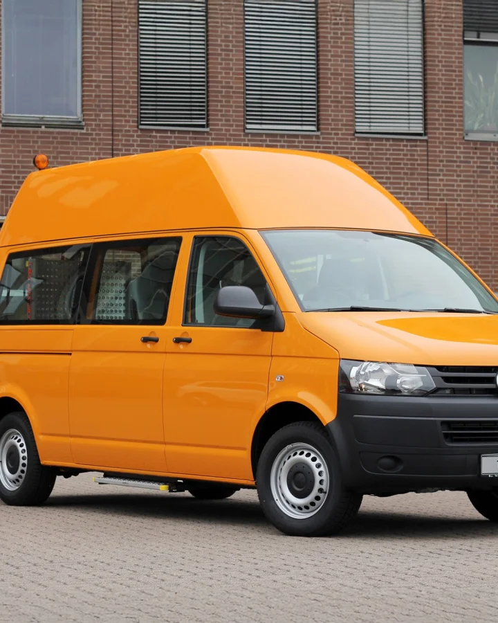 Photo of an orange high-roof VW Transporter parked in front of a brick building with large windows and garage doors.