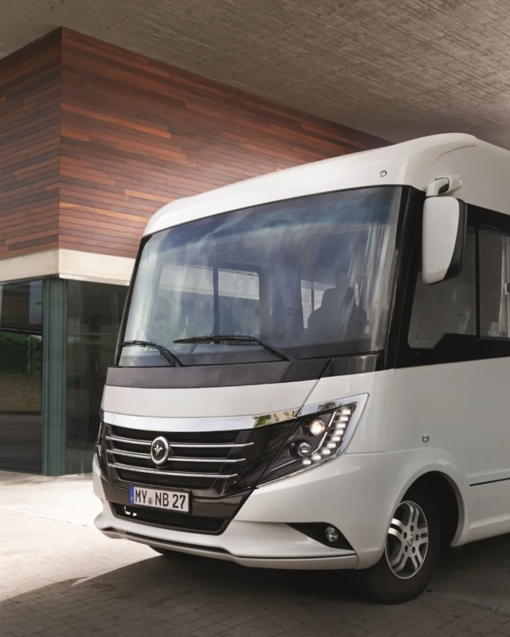 A modern white Niesmann+Bischoff motorhome parked in front of a contemporary building with wood and glass facade. The vehicle features high-quality fiberglass composite exterior panels manufactured by Fassmer for premium motorhomes.