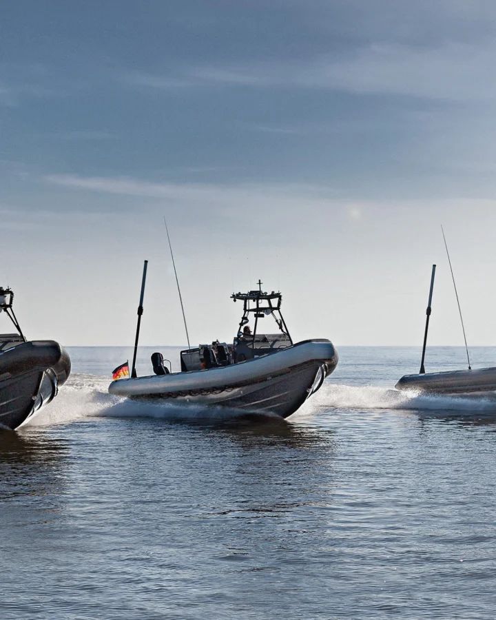 Three military RIB fast boats by Fassmer Defence at high-speed operation, navy vessel in background