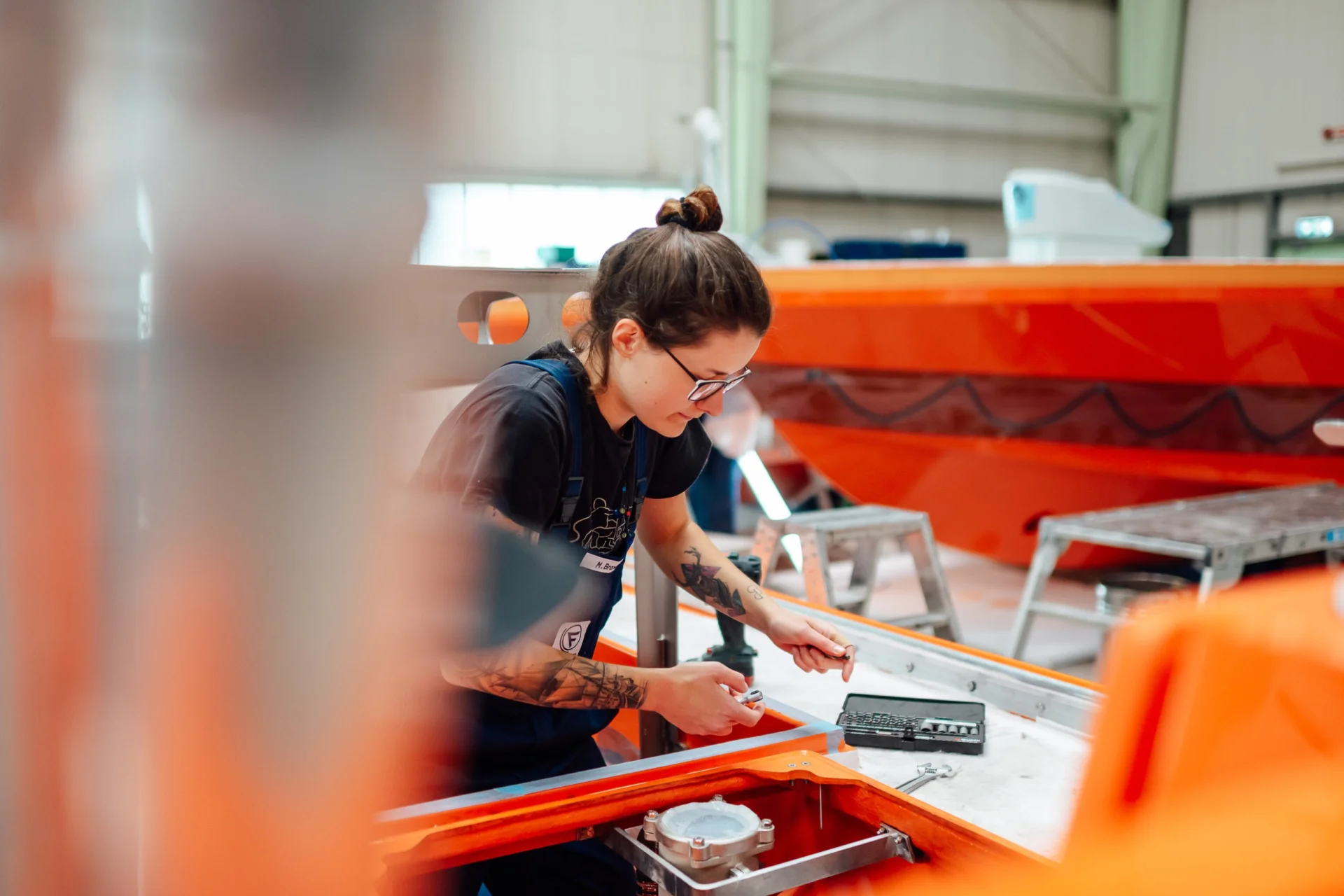 Trainee at Fassmer working in the shipyard: Young woman with glasses and tattoos assembling components on an orange boat hull. Hands-on training in shipbuilding.